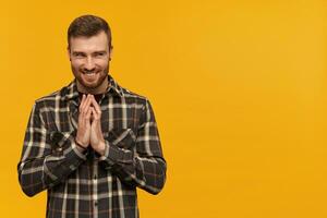 pernicioso arrogante jovem homem dentro xadrez camisa com barba fricção mãos e intrigante a mal plano sobre amarelo fundo olhando longe para a lado foto