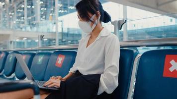 O viajante asiático da senhora de negócios usa máscara facial, sentado no banco, usa o laptop para trabalhar entre a espera pelo voo no terminal do aeroporto. viajante de negócios em uma pandemia covid, conceito de viagens de negócios.