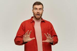 assustado chocado jovem homem dentro vermelho camisa com barba defendendo a partir de ameaças de mãos e gritando sobre branco fundo foto