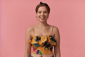 retrato do europeu atraente jovem mulher 20s com Castanho cabelo vestindo verão estilo colorida equipamento orelha argolas e bandana sorridente às você contra Rosa parede foto