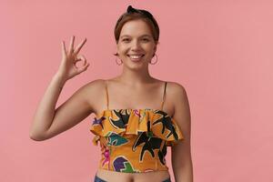 retrato do atraente jovem Castanho olhos mulher vestindo verão estilo colorida equipamento orelha argolas e Preto bandana olhando às você contra Rosa parede emoção feliz feliz sorridente às você confiante dedos OK foto