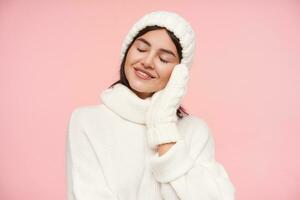 Boa olhando jovem satisfeito Castanho cabelos fêmea vestido dentro branco acolhedor roupas sorridente de bom grado com fechadas olhos e inclinado bochecha em elevado mão, posando sobre Rosa fundo foto