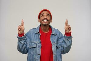 alegre jovem adorável Sombrio esfolado morena homem com barba cruzando dedos para Boa sorte e sorridente amplamente enquanto olhando para cima, posando sobre branco fundo foto