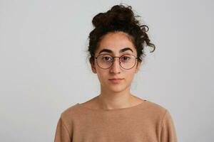 retrato do sério, adulto menina com Sombrio encaracolado cabelo pão. vestindo bege saltador e óculos. emoção conceito. assistindo às a Câmera. fechar acima isolado sobre branco fundo foto