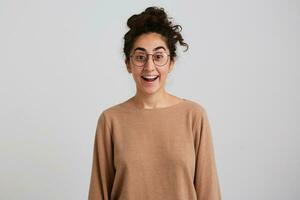 retrato do atraente, surpreso menina com Sombrio encaracolado cabelo pão. vestindo bege saltador e óculos. emoção conceito. animado sobre o que ela vê. assistindo às a Câmera isolado sobre branco fundo foto