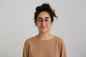 fofa olhando mulher, lindo menina com Sombrio encaracolado cabelo pão. vestindo bege saltador e óculos. emoção conceito. assistindo com sorrir às a Câmera isolado sobre branco fundo foto