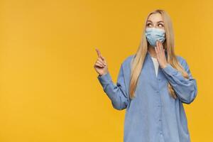 agradável olhando mulher, lindo menina com grandes loiro cabelo. vestindo azul camisa e médico face mascarar. assistindo às a Câmera e apontando para a esquerda às cópia de espaço, isolado sobre laranja fundo foto