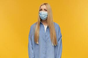 retrato do inseguro, adulto menina com loiro grandes cabelo. vestindo azul camisa e médico face mascarar. pessoas e emoção conceito. assistindo incrédulamente às a Câmera, isolado sobre laranja fundo foto