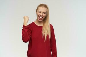 lindo menina com grandes loiro cabelo. vestindo vermelho suéter. pessoas e emoção conceito. assistindo às a Câmera, isolado sobre branco fundo, sorrisos amplamente e mostrar ganhar placa com mão foto