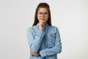 retrato do uma menina com óculos quem estão infeliz com o que é acontecendo, cruzado 1 mão, de outros 1 segurando a queixo 1 canto do lábios franzido, vestido dentro uma jeans camisa isolado em uma branco fundo foto