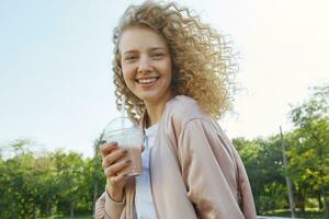 fechar acima do atraente jovem lindo menina Loiras com encaracolado cabelo anda em dentro a parque, bebendo uma coquetel, uma batido, vestindo uma Rosa Jaqueta sobre uma branco camiseta, sorridente e olhando para dentro a Câmera foto