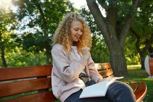 estudante, lindo cabelo loiro menina senta em uma parque banco, com uma bloco de anotações, parece em dela notas, pensa sobre tarefa, tentando para encontrar uma solução, segurando uma lápis dentro a boca e sorridente, parque fundo foto