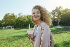 atraente jovem lindo menina Loiras com encaracolado cabelo anda em dentro a parque, vento ondas dela cabelo, bebidas uma coquetel, uma smoothie, usa uma Rosa jaqueta, sorridente, em pé lateralmente e olhando para dentro a Câmera foto