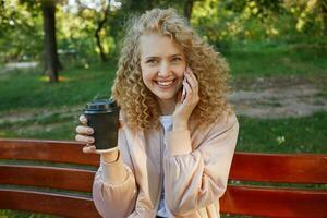 ao ar livre foto do jovem lindo menina Loiras senta em uma parque banco, bebendo café, Falando com alguém em telefone, sorridente, sente feliz, tem agradável conversação, parque fundo