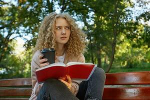 uma jovem lindo menina Loiras senta com dela pernas dobrado em uma parque banco, com café, notas, uma bloco de anotações, parece ausente, pensa sobre algo, tentando para encontrar uma solução, mordeu dela mais baixo lábio foto