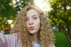 lindo atraente menina Loiras com encaracolado cabelo e azul encantador olhos, enviando sopro beijo com beicinho lábios olhando às Câmera, fazer selfie, natureza fundo foto