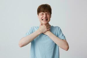 retrato do sem esperança estressado jovem homem com fechadas olhos e suspensórios em dentes desgasta azul t camisa estrangula ele mesmo com ambos mãos isolado sobre branco fundo foto