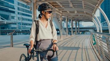 mulher asiática empresária usar óculos de sol ir trabalhar no escritório andar e sorrir olhar ao redor segurar o carrinho de bicicleta ao redor do edifício em uma rua da cidade. bicicleta pendulares, comutar de bicicleta, conceito de viajante de negócios.