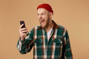 retrato do Bravo jovem homem gritando em dele Móvel telefone sobre bege fundo. louco irritado barbudo jovem homem dentro xadrez camisa segurando e gritando em célula telefone foto