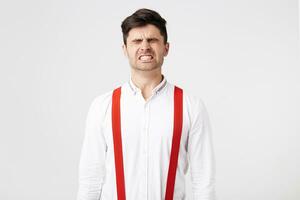 uma homem vestido dentro uma branco camisa e vermelho suspensórios, parafuso acima dele olhos, mostrando, apertando branco dentes Como E se ele teve levantado alguma coisa pesado, a insuportável fardo, forte dor, isolada sobre branco fundo foto