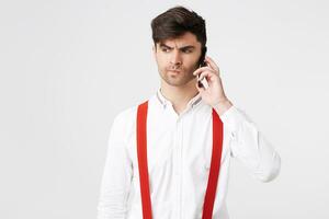 cabelos escuros homem falando em uma Móvel telefone, desgasta uma camisa e vermelho suspensórios parece desapontado descontente parece para a lado franzido dele lábios, um desagradável Telefone conversação trouxe em triste pensamentos foto