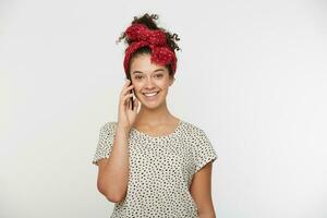 alegre jovem menina brilhando a partir de felicidade, falando com dela querida namorado em telefone, olhando feliz e alegre, mantém inteligente telefone dentro mão, vestido branco camiseta com pontos, branco fundo foto