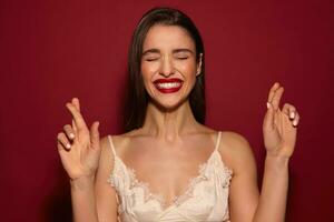 retrato do jovem encantador grandes cabelos morena senhora com vermelho lábios levantando mãos com cruzado dedos e sorridente amplamente enquanto em pé sobre Borgonha fundo foto