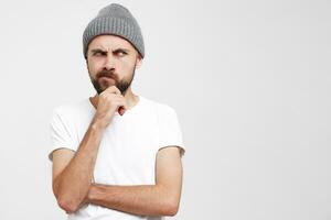 jovem europeu homem acha, elevado mão para face, toques dele barba, parece insatisfeito, 1 canto do a lábios franzido, vestido dentro uma em branco branco camiseta e cinzento chapéu, isolado sobre branco fundo foto