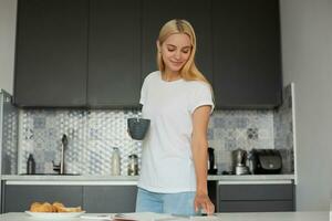 Loiras menina em pé perto a mesa, sorridente, tendo café da manhã, planejamento dela dia, mantém uma grande cinzento xícara, conduz dele dedo através a telefone tela, Verificações correspondência, cozinha fundo foto