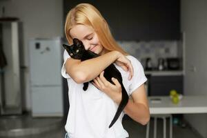 fechar acima do jovem Loiras menina às a cozinha detém em mãos, carícias, acariciando, abraços Preto pequeno gato, fofa ternamente sorridente, olhos fechado, vestido dentro uma doméstico branco camiseta, cozinha fundo foto