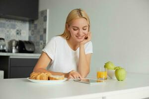 adorável concurso atraente mulher senta às a mesa, sorridente, parece às tábua Preto tela, lendo mensagem, em a mesa - uma vidro do suco, maçãs, pães, vestido dentro doméstico camiseta, cozinha fundo foto