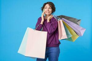 horizontal tiro do bonita jovem curto cabelos encaracolado mulher tendo telefone conversação enquanto caminhando com papel bolsas sobre azul fundo, vestido dentro casual roupas foto