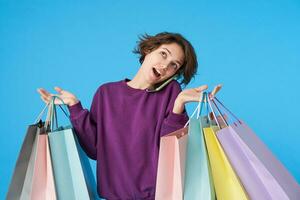 interior tiro do jovem bonita encaracolado morena mulher com curto corte de cabelo fazer ligar com dela Smartphone enquanto compras, guardando papel bolsas dentro elevado mãos, isolado sobre azul fundo foto