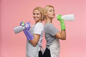 positivo jovem atraente cabeça branca limpeza senhoras vestindo borracha luvas enquanto limpeza casa e segurando spray garrafas, em pé sobre Rosa fundo foto