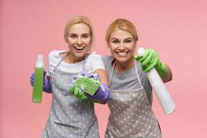 feliz jovem Loiras donas de casa dentro borracha luvas olhando alegremente às Câmera com Largo sorrir e guardando garrafas com detergentes, em pé posando sobre Rosa fundo foto