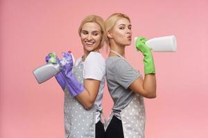 interior foto do jovem Loiras fêmeas segurando spray garrafas enquanto inclinado em cada outro, vestindo casual Camisetas e polca ponto avental enquanto posando sobre Rosa fundo