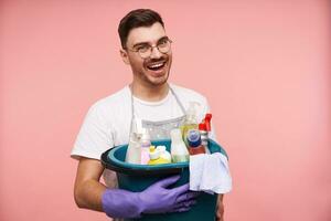 interior tiro do feliz jovem barba por fazer morena homem dentro uniforme preparando para Primavera limpeza e segurando família produtos químicos enquanto posando sobre Rosa fundo foto