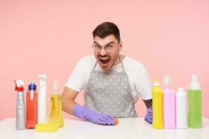 irritado jovem Sombrio cabelos barba por fazer masculino com curto corte de cabelo gritando com Largo boca aberto enquanto sentado sobre Rosa fundo com garrafas do detergentes foto