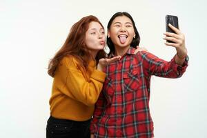 retrato do ásia e caucasiano amigos. vestindo amarelo suéter e xadrez camisa. enviando ar beijo e mostrando língua, fazer selfie em Smartphone. ficar de pé isolado sobre branco fundo foto