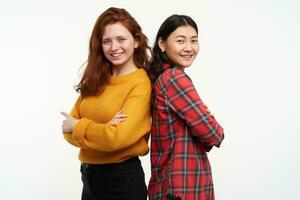 dois jovem mulheres amigos. vestindo amarelo suéter e xadrez camisa. em pé costas para costas com braços cruzado. pessoas e estilo de vida conceito. assistindo às a Câmera, isolado sobre branco fundo foto