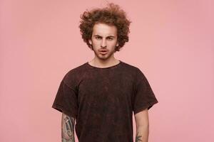 legal olhando homem, bonito cara com tatuagens, morena encaracolado cabelo e barba. vestindo Sombrio vermelho camiseta. emoção e pessoas conceito. assistindo sério às a Câmera isolado sobre pastel Rosa fundo foto