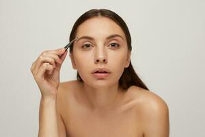 fechar acima do uma lindo mulher Como em pé perto uma espelho. jovem bonita menina cuidados para a face, arranca sobrancelhas com pinças, isolado em branco fundo. cabeça e ombros foto