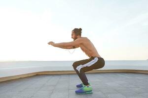 retrato do jovem barbudo homem com em forma Atlético corpo fazendo alongamento exercícios, ouvindo música em fones de ouvido, tem muscular corpo forma. foto