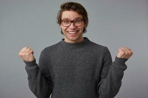 jovem feliz cara com óculos desgasta dentro cinzento suéter, carrinhos sobre cinzento fundo. amplamente sorrisos e aperta punhos, Ganhou a e sente ótimo. foto