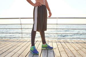 foto do pernas do desportivo homem, desgasta é colorida tênis e listrado shorts. ficar de pé às a à beira-mar.