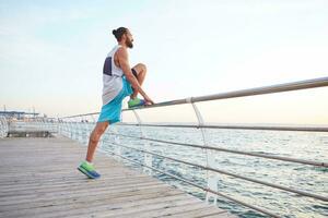 jovem barbudo atraente cara fazendo manhã exercícios de a mar, aquecer depois de correr, alongamento para pernas, conduz saudável ativo estilo de vida. ginástica masculino modelo. foto