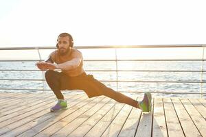 jovem barbudo atraente cara fazendo manhã exercícios de a mar, ouvindo favorito música em fones de ouvido, aquecer depois de correr, alongamento para pernas, conduz saudável ativo estilo de vida. ginástica masculino modelo. foto