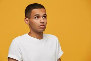 atraente jovem homem parece a parte, de lado acima intrigado, tentando para lembrar alguma coisa importante, considerado. Serra alguma coisa fascinante dentro a céu. desgasta branco casual camiseta, em uma amarelo fundo foto