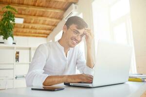 retrato do feliz confiante jovem homem de negocios desgasta branco camisa e óculos usando computador portátil e Smartphone sentado às a mesa, trabalhando dentro escritório e rindo foto