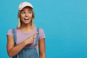 fechar-se do feliz espantado Loiras jovem mulher desgasta Rosa boné, tolet t camisa e jeans macacão parece surpreso e pontos para a lado isolado sobre azul fundo foto